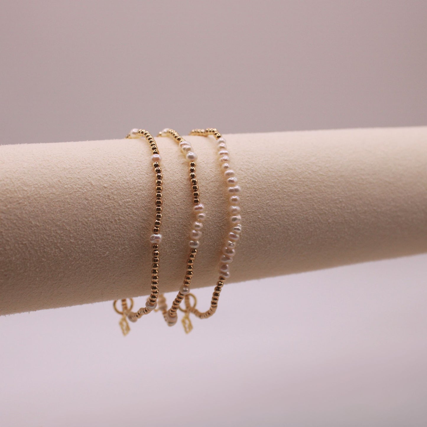 A trio of bracelets displayed on a cylindrical stand, featuring freshwater pearls and metallic beads with a small charm.