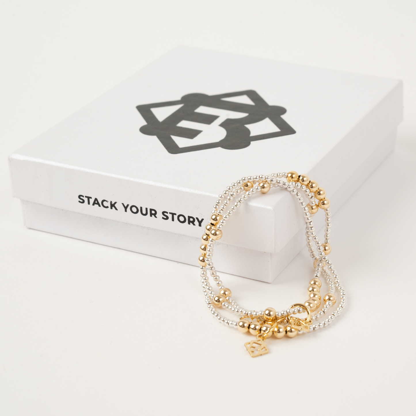 Stack of gold and silver bracelets on a white box with 'Stack Your Story' branding.