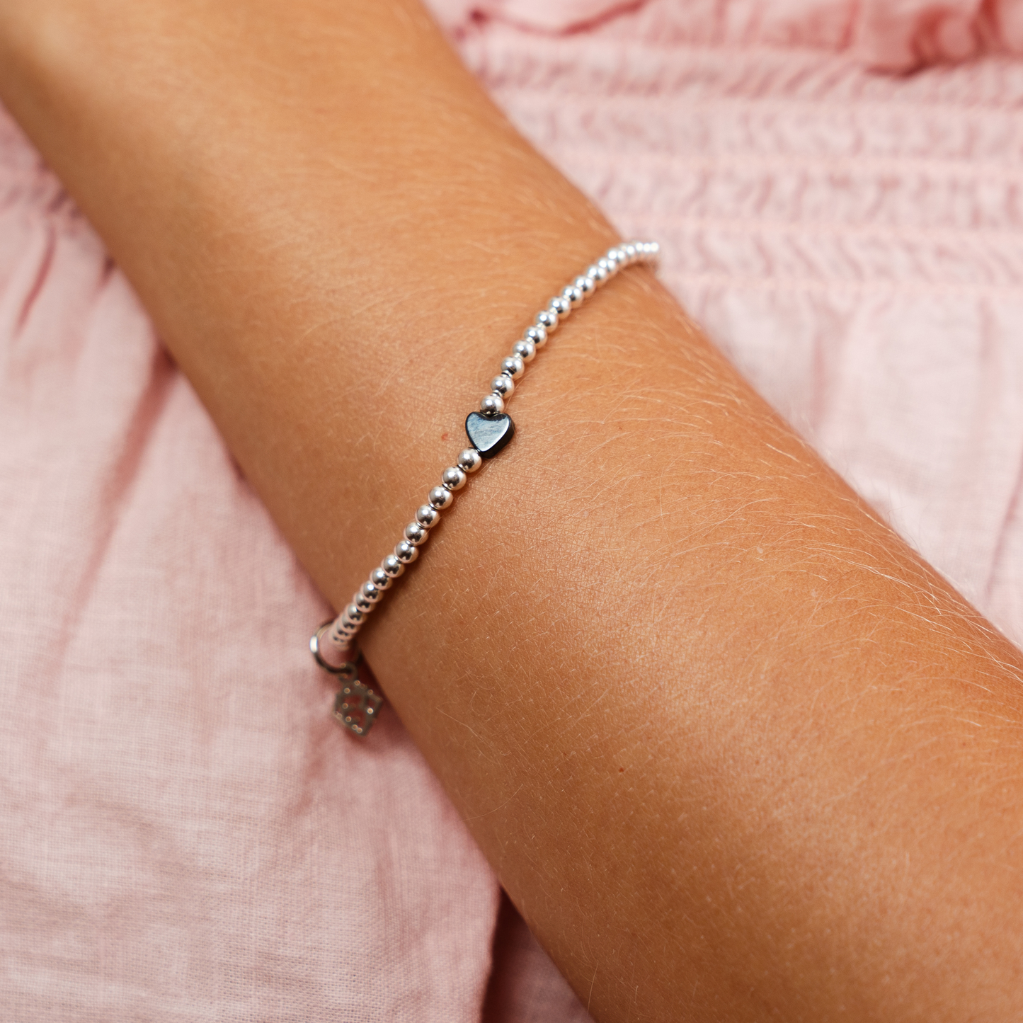 Silver bracelet with a heart charm on a wrist against a pink background
