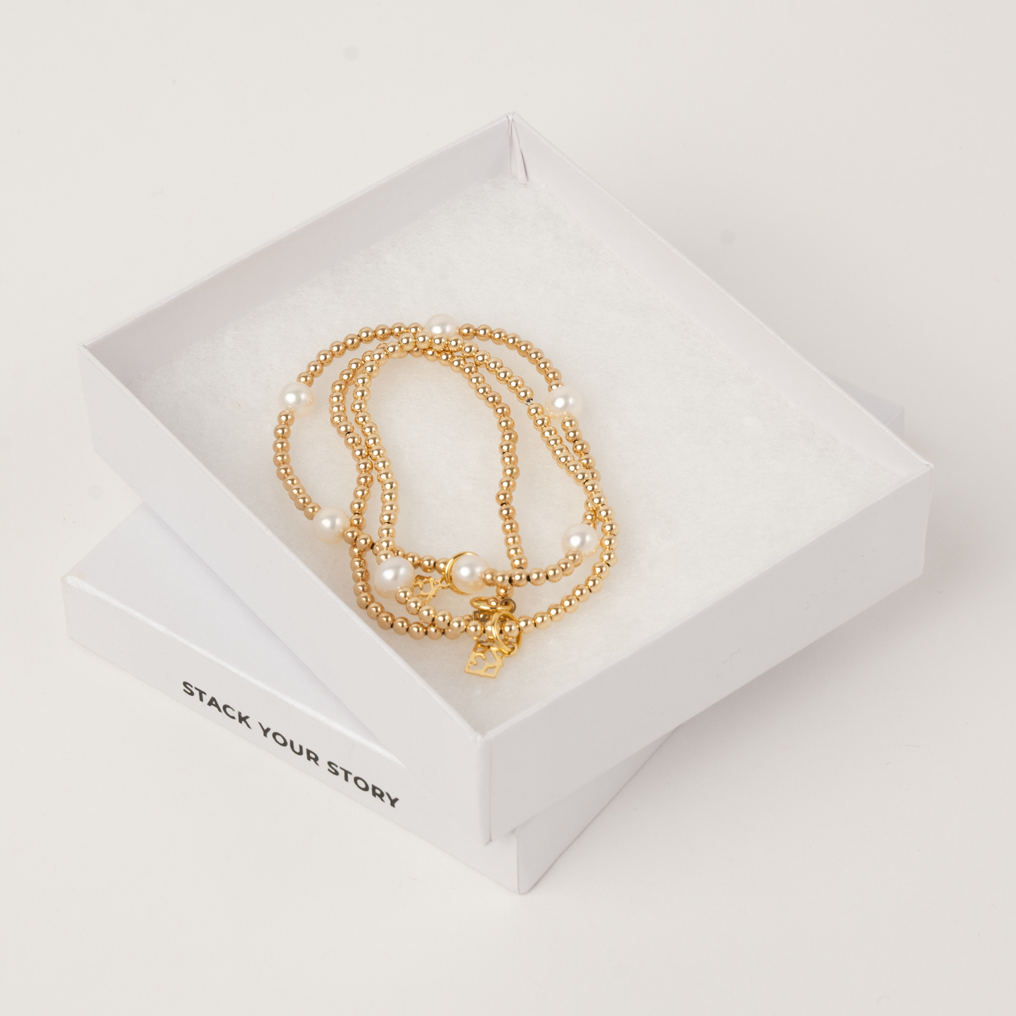 Gold beaded bracelets on a white box with 'Stack Your Story' text.