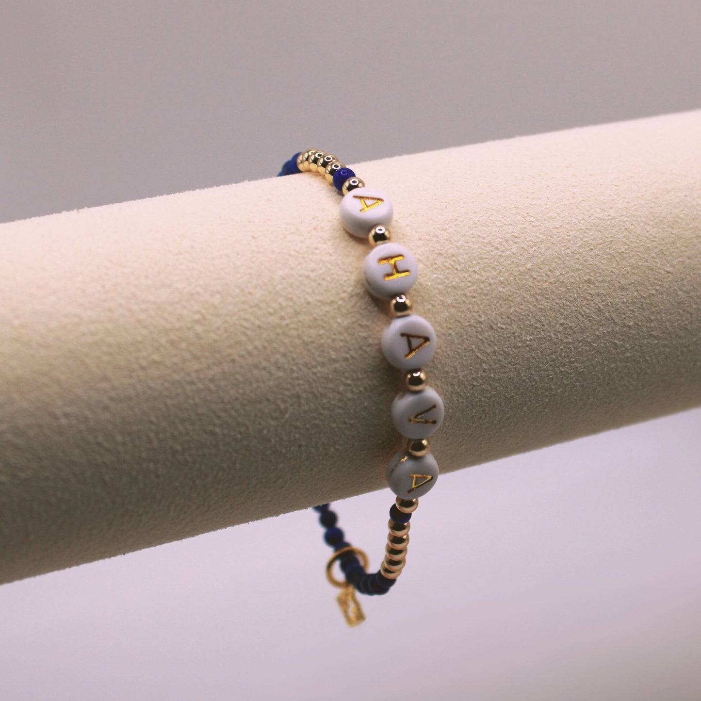 A bracelet featuring gold-colored beads and gray beads with the word 'AHAVA' in Hebrew, displayed on a cylindrical stand.