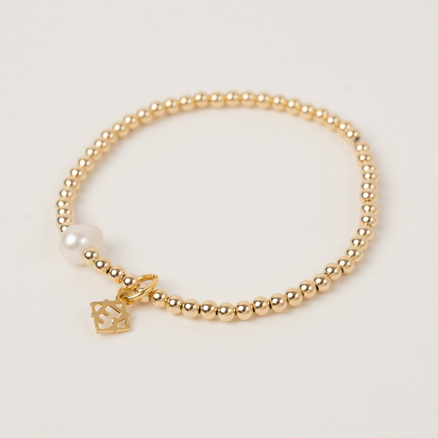Gold beaded bracelet with a pearl and square charm on a light gray background