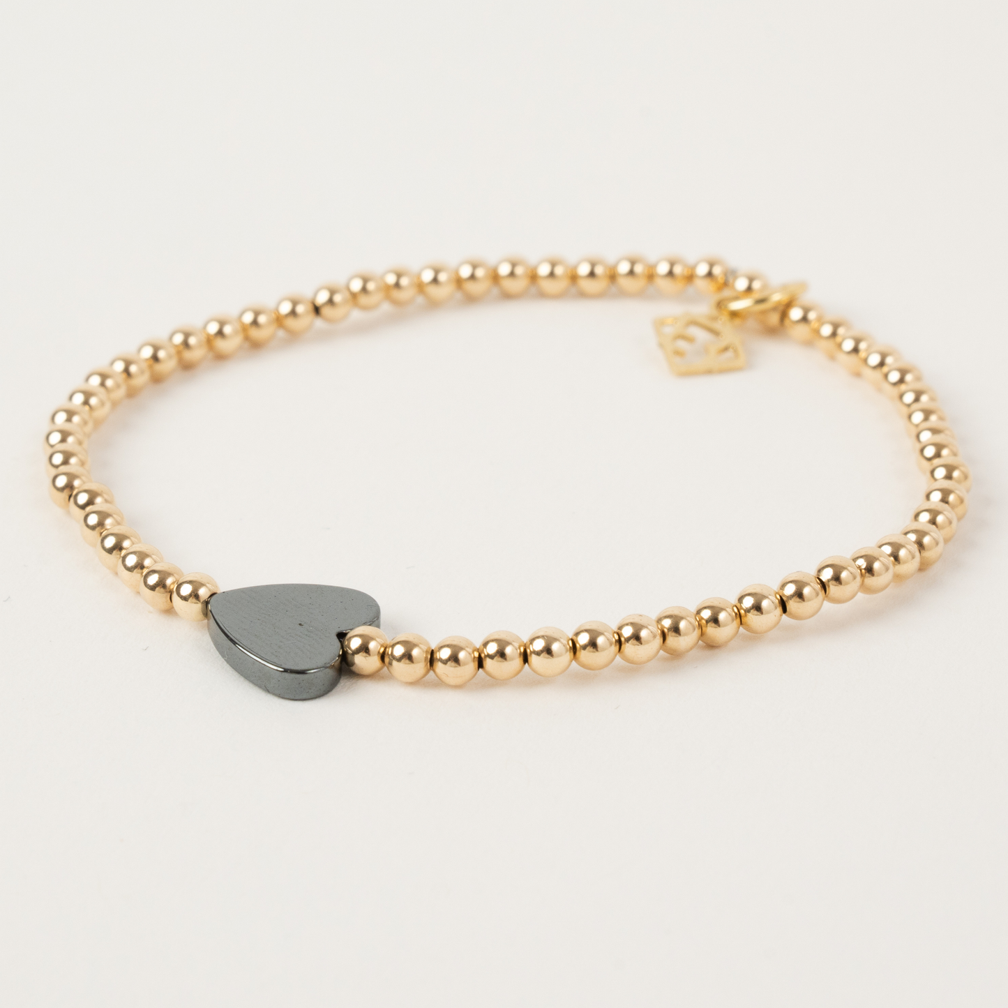 Gold beaded bracelet with a heart-shaped black charm on a light gray background