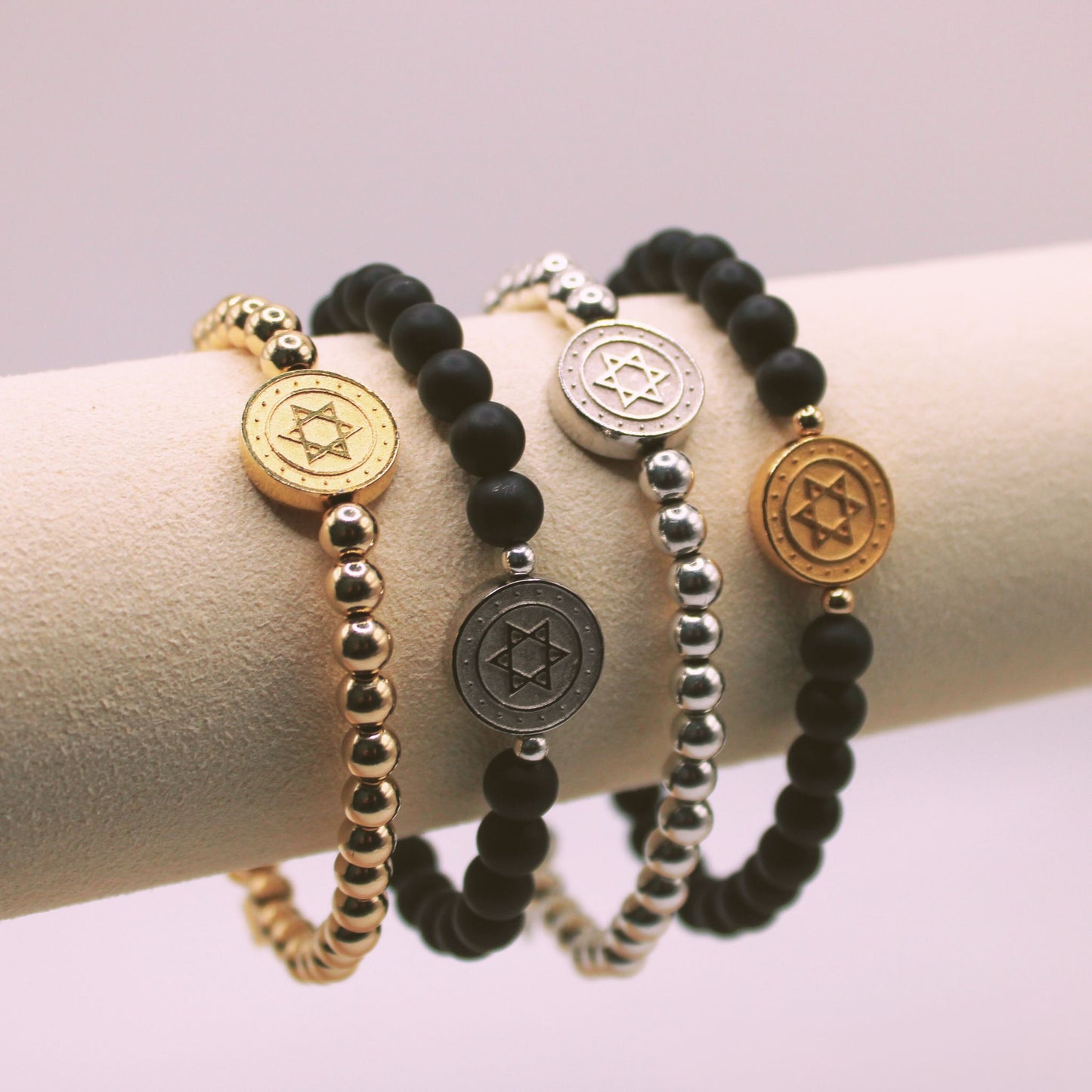 A collection of four Star of David bracelets displayed on a cylindrical object, featuring variations in gold, silver, matte black, and lapis colors with bead details.