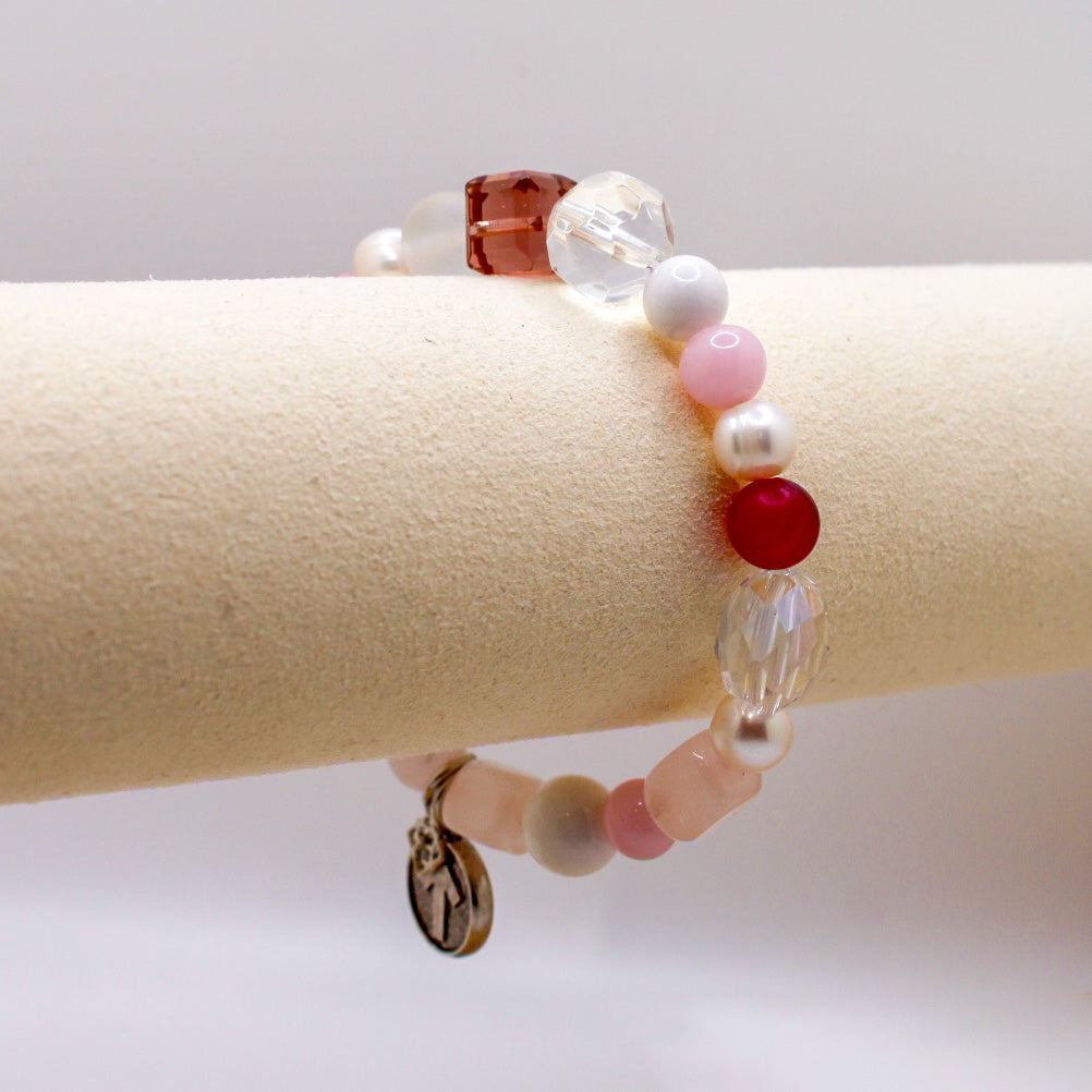 A pink and white beaded bracelet with semi-precious stones and pearls, featuring a signature arrow charm and an iconic dangle charm, displayed on a beige cylindrical object.