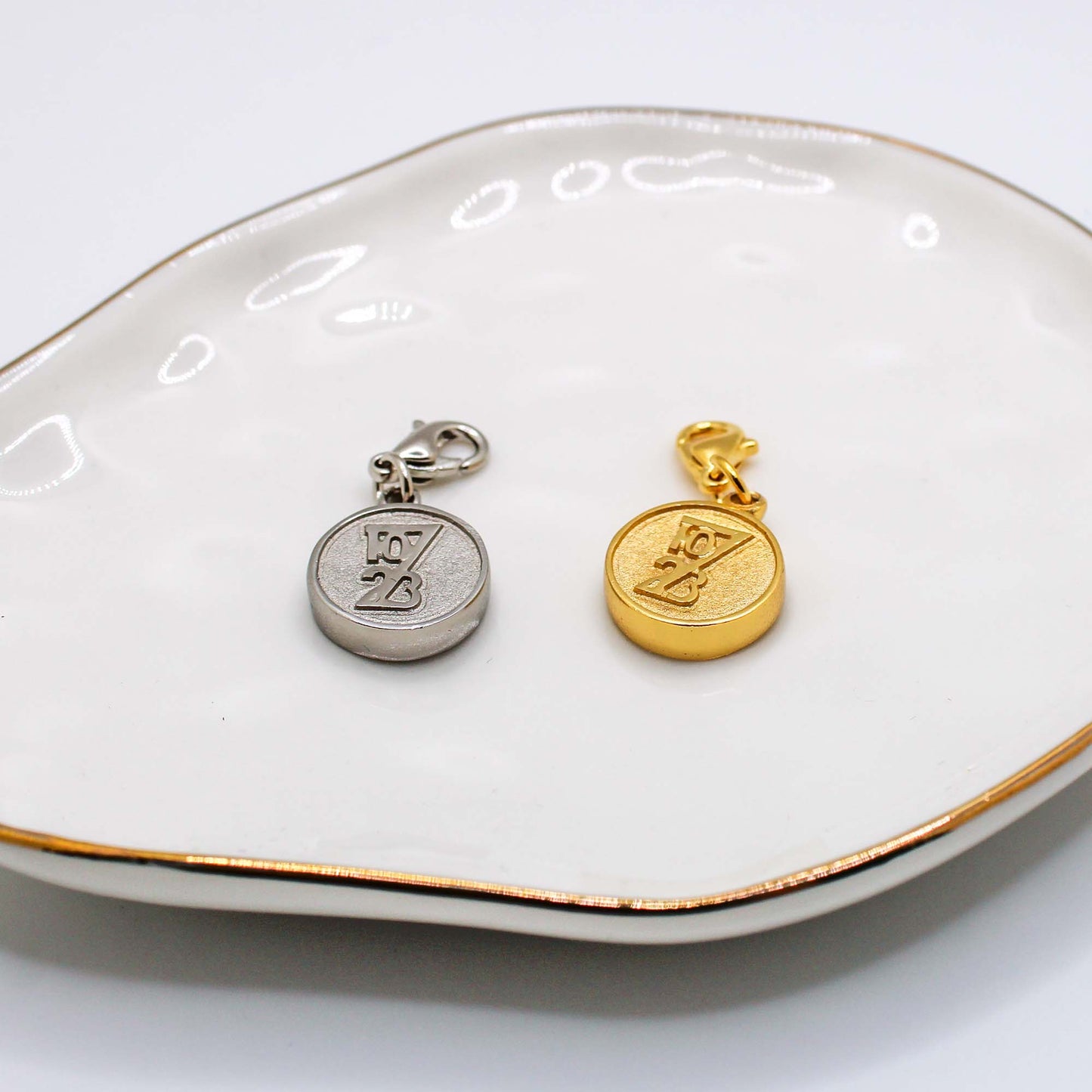 Two charity pendants on a white dish, one silver and one gold, each with a different design.