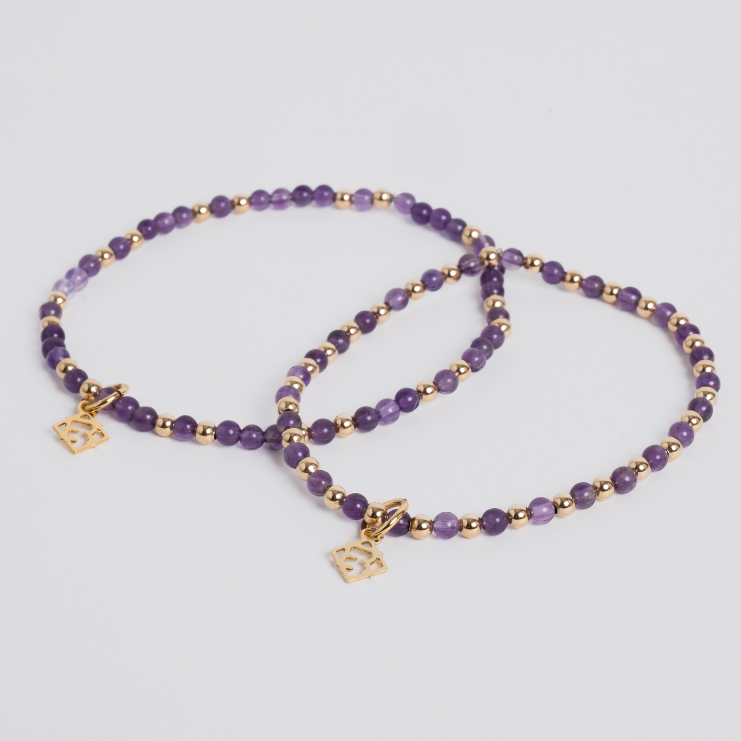 Two beaded stretch bracelets with purple gemstone beads, small gold spacer beads, and gold-toned square charms, displayed on a light gray background.