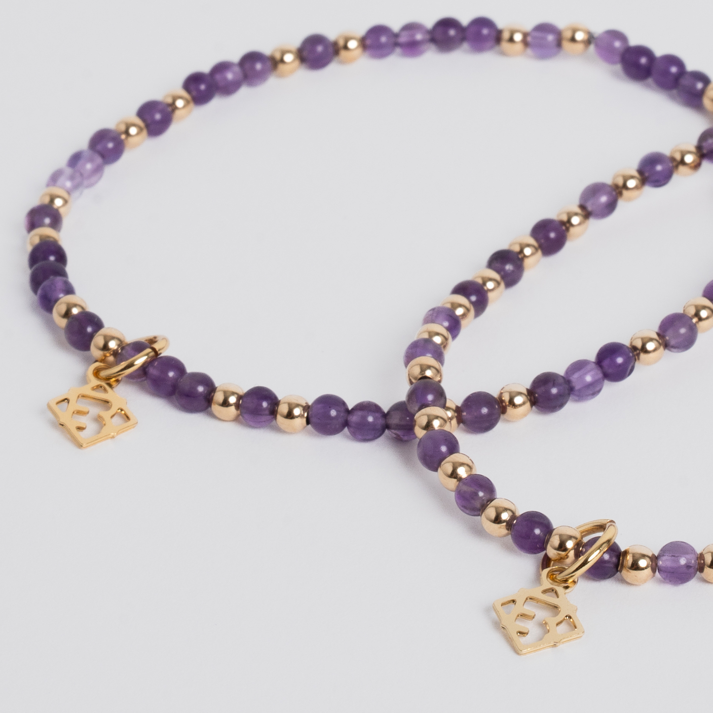 Purple and gold beaded necklace with two small square gold charms, photographed on a light gray background.