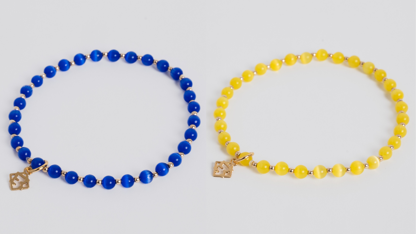 Two beaded bracelets displayed side by side on a light gray background. The left bracelet is made of round blue beads with small gold spacer beads and a gold charm.