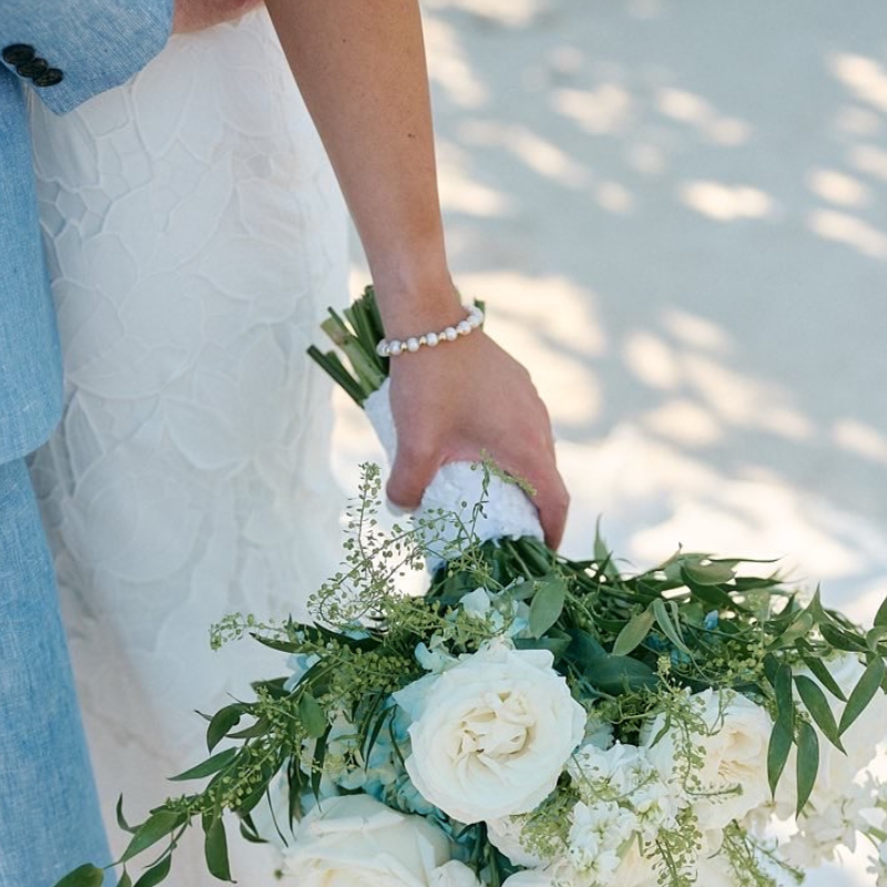 Person holding a bouquet of white flowers with green leaves on a sandy background