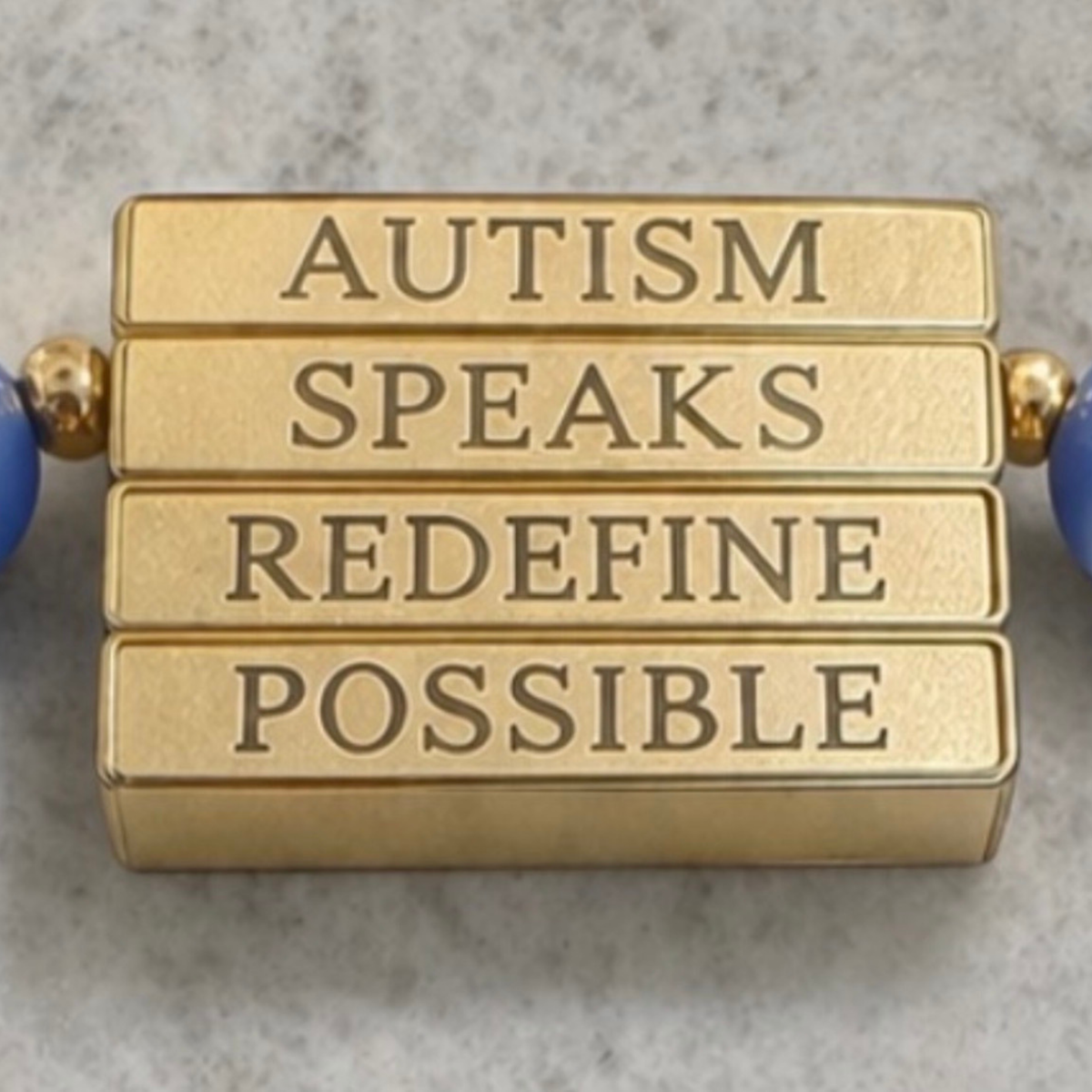 Close-up of the Autism Speaks Gold Bracelet with blue cat’s-eye gemstones and a rotating mantra bar engraved with Autism Speaks Redefine Possible