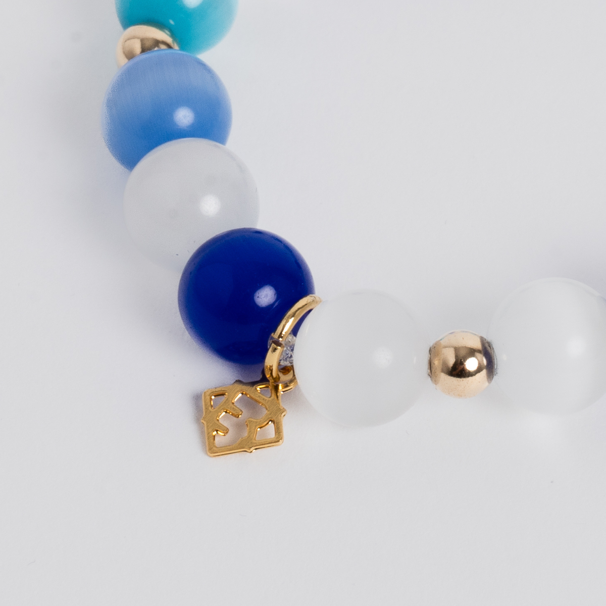 Close-up of the Prism Maxi Multi Blues Bracelet with white, royal blue, sky blue and turquoise cat’s-eye beads and a golden ER charm