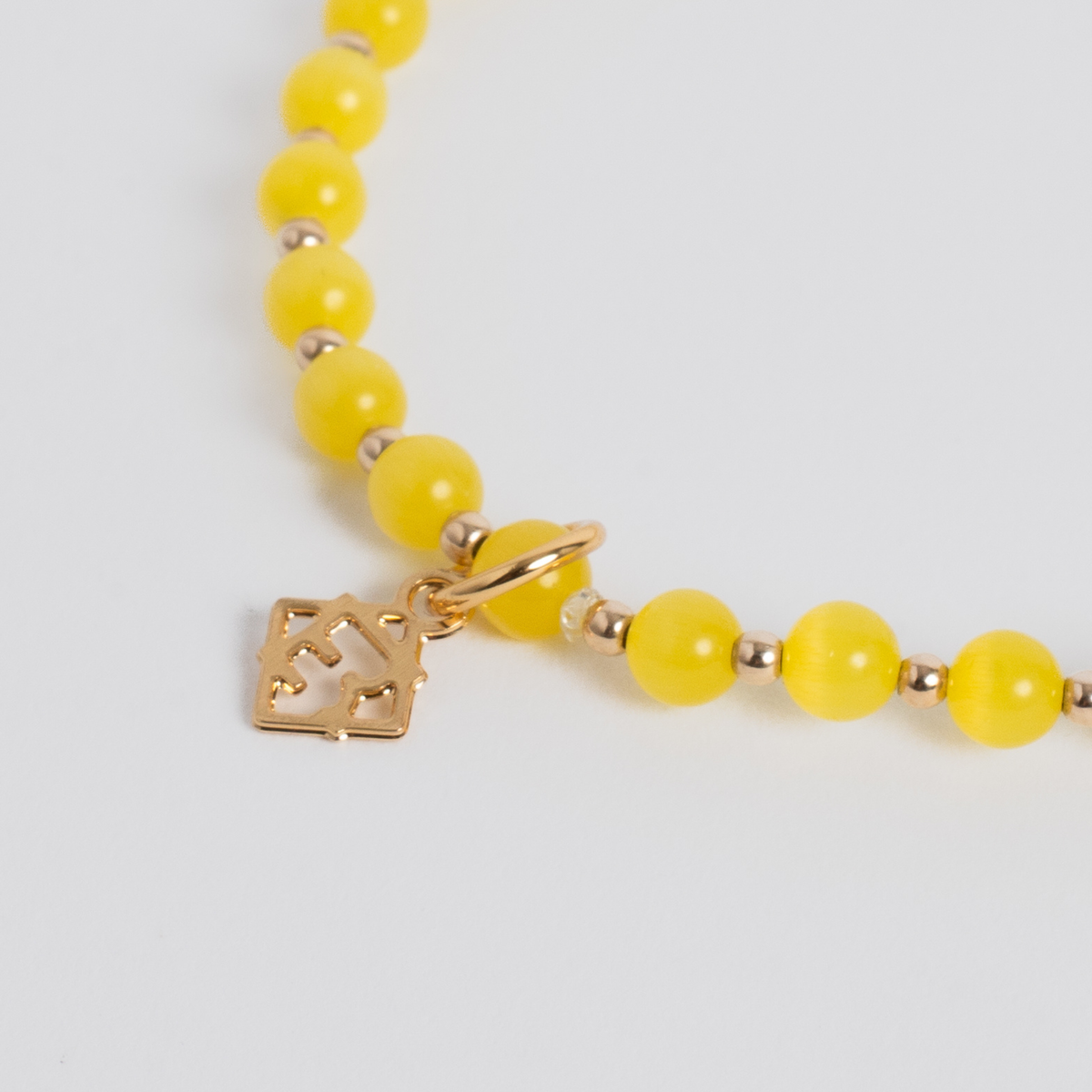 A close-up of a yellow beaded necklace with small metallic spacer beads and a gold-tone square charm featuring a mountain-like cutout design, shown on a light gray background.